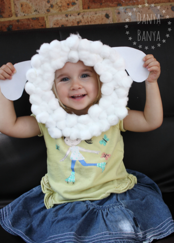Sheep mask made with a paper plate and cotton balls held by a little girl in yellow and a blue skirt.